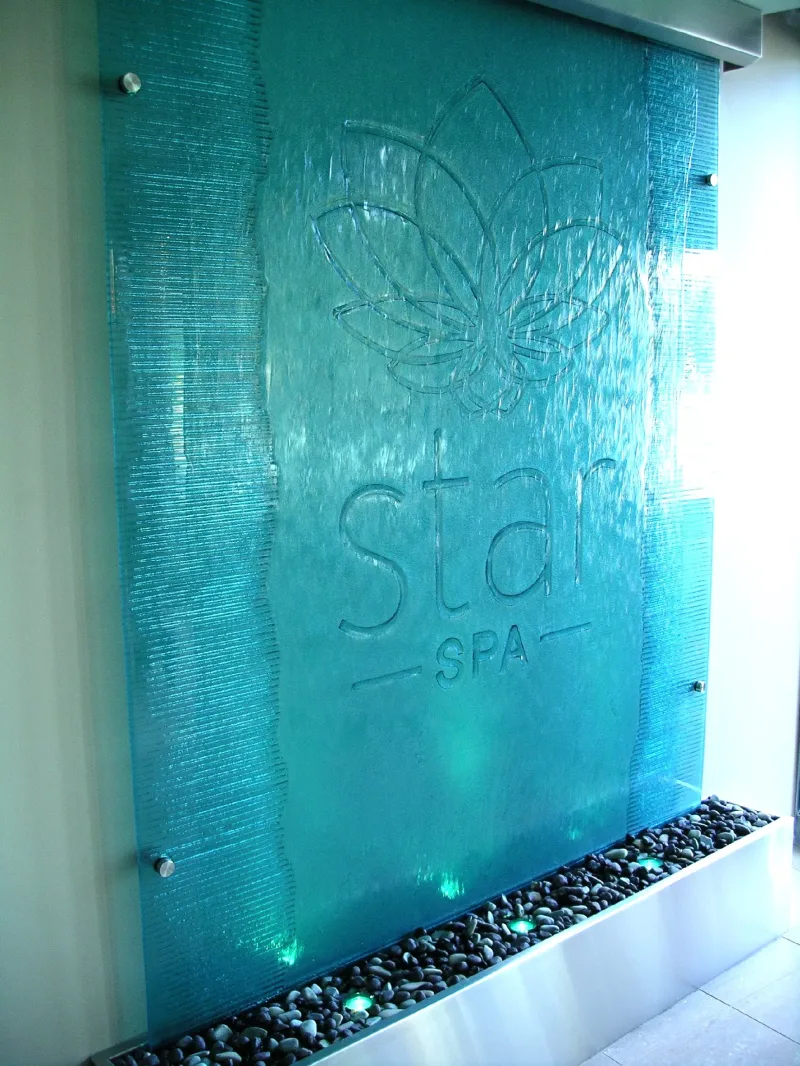 Teal textured glass water wall with an etched spa logo above a pebble trough with LED lights